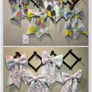 Puede incluir: Colección de lazos acolchados hechos a mano en varios colores y patrones, exhibidos en una pared. Los lazos están sujetos a marcos de madera decorativos. Algunos tienen un diseño de patchwork, otros son blancos con patrones de estrellas rosas.