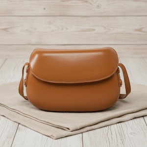 May include: A brown, rounded crossbody bag with a flap closure and a long shoulder strap. The bag is made of a smooth material and sits on a beige cloth. The background is a light-colored wooden surface.