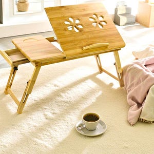 May include: A bamboo laptop desk with a tilting surface and a small drawer. The desk has two flower-shaped cutouts and a small lip to prevent items from sliding off. A cup of coffee sits on a saucer nearby.