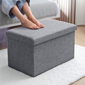 May include: A grey storage ottoman with a hinged lid. The rectangular ottoman is made of a textured fabric and sits on a white rug. The ottoman is a neutral colour and can be used for storage or as a footrest.