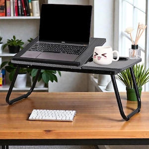 May include: A grey laptop desk with black metal legs, holding a laptop and a white mug with a face design. The desk has a built-in cup holder and a slot for a laptop. A wireless keyboard sits on the wooden surface below.