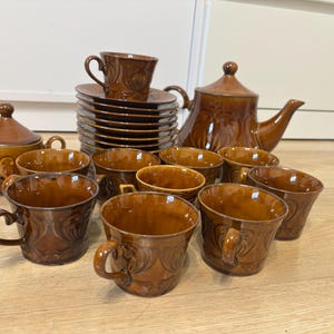 May include: A brown ceramic tea set with a teapot, sugar bowl, cups, saucers, and a cup on top of a stack of saucers. The set has a textured design and a glossy finish. The cups have handles.