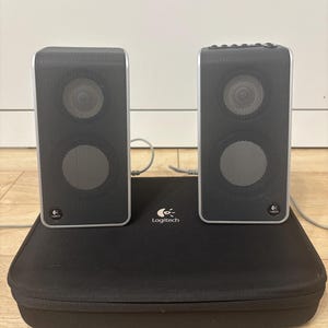 Może przedstawiać: Dwa czarne głośniki komputerowe Logitech ze srebrnymi obramowaniami, każdy z dwoma okrągłymi otworami głośnikowymi. Stoją na czarnej, prostokątnej obudowie z logo Logitech. Głośniki połączone są szarym kablem.