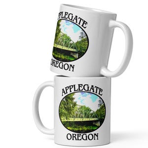 Può includere: Due tazze di ceramica bianca impilate l'una sull'altra. Ogni tazza presenta un'immagine circolare di un ponte verde su un fiume, circondato da alberi, con le parole "Applegate Oregon" stampate intorno all'immagine.