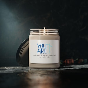 May include: A lit candle in a glass jar with a silver lid. The label reads "YOU ARE enough" in blue text. Below the main text is the phrase "Actually, most of the time you're a little too much dial it back a smidge."