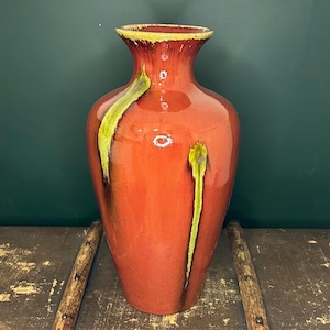 May include: A tall, glossy red vase with a flared rim and a rounded body. The vase features vertical streaks of yellow-green paint. The rim is a darker shade of red with a yellow-brown edge. The vase is a decorative home accent.