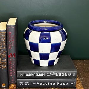 May include: A blue and white checked ceramic vase with a blue rim, resting on a stack of books. The books include titles such as "Texas Hold 'Em" and "Thursday Murder Club". The vase is a decorative item.