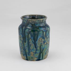 May include: A ceramic vase with a cylindrical shape and a slightly flared rim. The vase features a glaze with a blend of blue, teal, and gold tones, creating a dripped effect. The overall aesthetic is rustic and artistic.