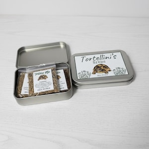 May include: Two rectangular silver metal tins. One tin is open, containing clear plastic bags filled with dried plant matter and labels reading Tortoise Mix and Zeus World. The other tin is closed with a label featuring a tortoise illustration and the text Tortellini's Seeds Zeus World.