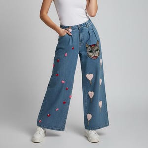 May include: Light blue denim wide-leg trousers with embroidered cat face and heart details. The trousers feature a high waist and are paired with a white vest top and white trainers. The cat face is on the right leg, and the hearts are on both legs.