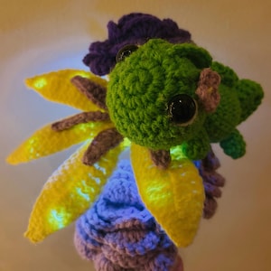 May include: A handmade crochet flower with a green head, large black eyes, and yellow petals. The petals are outlined with brown and have a glowing effect. The flower has a purple and blue base and a purple flower on top.