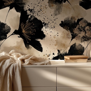 May include: A white dresser featuring a draped cream-colored fabric, a stack of two books, and a beige vase with dried stems. The wall behind displays a wallpaper pattern of large, abstract black flowers with gold accents and ink splatters.