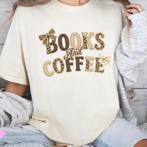 Può includere: T-shirt color avorio con la scritta "BOOKS And COFFEE" in lettere marroni, beige e con stampa leopardata. La maglietta ha una vestibilità ampia e maniche corte. Grafiche a fiocco sopra e sotto il testo.