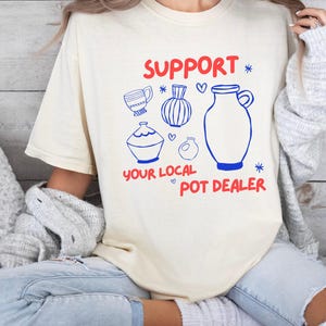 Puede incluir: Camiseta color crema con el texto "SUPPORT YOUR LOCAL POT DEALER" en rojo y azul. La camiseta presenta dibujos lineales azules de varios objetos de cerámica, incluyendo una taza, jarrones y una jarra.