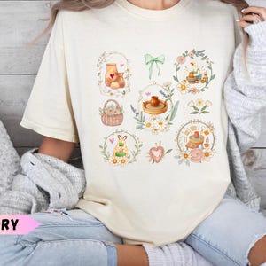 May include: An ivory short-sleeve t-shirt featuring a central graphic print of various watercolour-style illustrations including pottery, a basket of flowers, a green bow, a duck, and floral wreaths arranged in a grid-like pattern on the front.