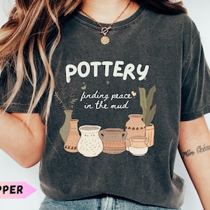 May include: Dark gray t-shirt with the word "POTTERY" in white, along with the phrase "finding peace in the mud." Below the text are illustrations of various pottery pieces and plants. The shirt is a relaxed fit.
