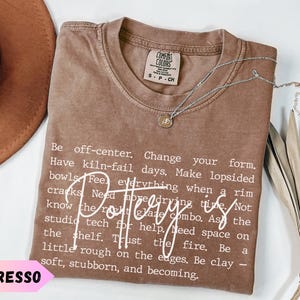 May include: Espresso-coloured t-shirt with white text about pottery. The shirt has a crew neck and short sleeves. A silver necklace with a small pendant rests on the shirt. A brown hat is visible in the background.