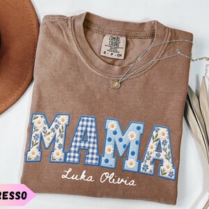 Puede incluir: Una camiseta color espresso con la palabra MAMA en letras grandes con borde azul. Las letras tienen estampados florales y de cuadros vichy. Debajo, los nombres Luka Olivia están bordados en letra blanca. Se ve un collar de plata.