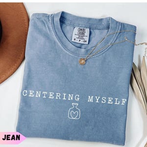 May include: A blue jean colored t-shirt with a crew neckline and a Comfort Colors label. The front features the white text "CENTERING MYSELF" above a simple white line drawing of a vase with a heart shape in the center of the vase outline.