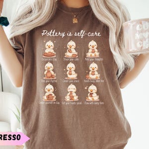 以下が含まれることがあります： 「Pottery is self-care」の文字とアヒルのイラストが描かれたエスプレッソカラーのTシャツ。各アヒルは陶芸に関連する活動に従事しています。シャツにはピンクのバナーで「ESPRESSO」の文字も入っています。