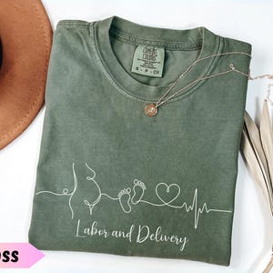 May include: A moss green t-shirt with the words "Labor and Delivery" in white script. The shirt features a white line drawing of a pregnant belly, footprints, a heart, and a heartbeat line. A gold necklace rests on the shirt.
