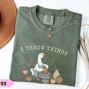 Może przedstawiać: Mchowo-zielony t-shirt z krótkim rękawem z grafiką przedstawiającą białą gęś w niebieskim fartuchu pracującą przy kole garncarskim. Nad gęsią znajduje się napis "I THROW THINGS". Koszulka posiada metkę marki Comfort Colors przy kołnierzu.