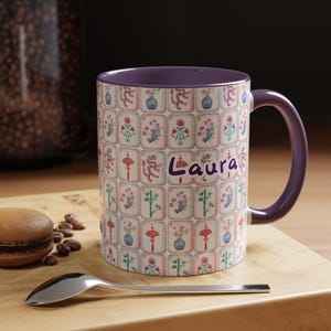 May include: A ceramic mug with a purple interior and handle, featuring a repeating pattern of floral and dragon designs in pink, blue, and red. The name "Laura" is written in purple script on the side. The mug sits on a wooden surface.