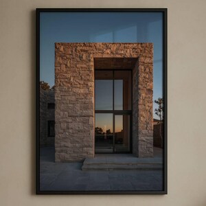 May include: A framed print of a building entrance with a stone facade and glass doors. The artwork is displayed above a bed with tan pillows and a cream-colored duvet. A black lamp sits on a nightstand.