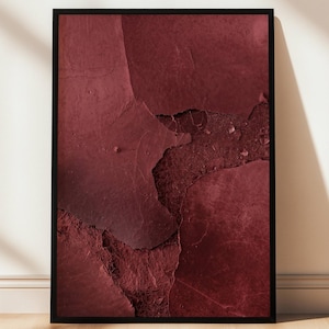 May include: A framed abstract art print featuring a close-up of a textured surface in shades of deep red and burgundy. The artwork has a cracked, weathered appearance, with a black frame. The print is leaning against a white wall on a wooden floor.