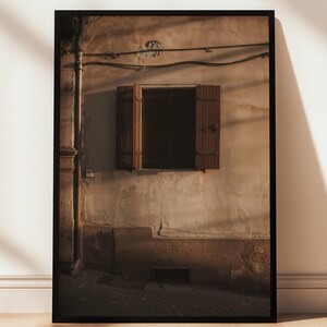 May include: Framed photograph of a weathered building facade with a window and closed wooden shutters. The wall is a mix of beige and brown tones, with shadows cast across the surface. The image is displayed on a wooden floor.