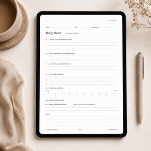 May include: A black tablet displaying a "Daily Reset" journal page with prompts for reflection. A beige pen rests beside the tablet. The journal prompts include questions about thoughts, priorities, and emotions. A cup of coffee and a beige cloth are in the background.