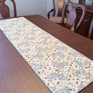 May include: A rectangular table runner with a butterfly pattern. The runner features a light blue and cream background with repeating butterfly and floral motifs. The runner is placed on a dark brown wooden table.