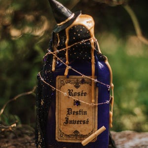 Peut inclure: Une bouteille de potion bleu foncé avec un chapeau de sorcière, des accents dorés et une étiquette indiquant "Rosée du Destin Inversé". La bouteille est décorée d'un tissu noir, de ficelle et de petits cristaux. Un parchemin roulé repose près de la base.