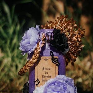 May include: A decorative purple bottle with a gold braided accent, adorned with a black flower, gold floral accents, and light purple and white flowers. The bottle has a label with French text and musical notes.