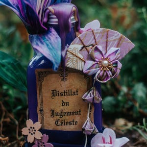 Op de afbeelding: Een decoratieve paarse fles met het opschrift "Distillat du Jugement Celeste". De fles is versierd met paarse en blauwe bloemaccenten, een waaiervormig ornament en een bloem. De fles is ongeveer 15 cm hoog.