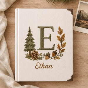 May include: A white fabric-covered book featuring an embroidered green letter E, a pine tree, a pinecone, and acorns. The name Ethan is embroidered in brown script below the letter. The book has decorative metal corner protectors on the cover.