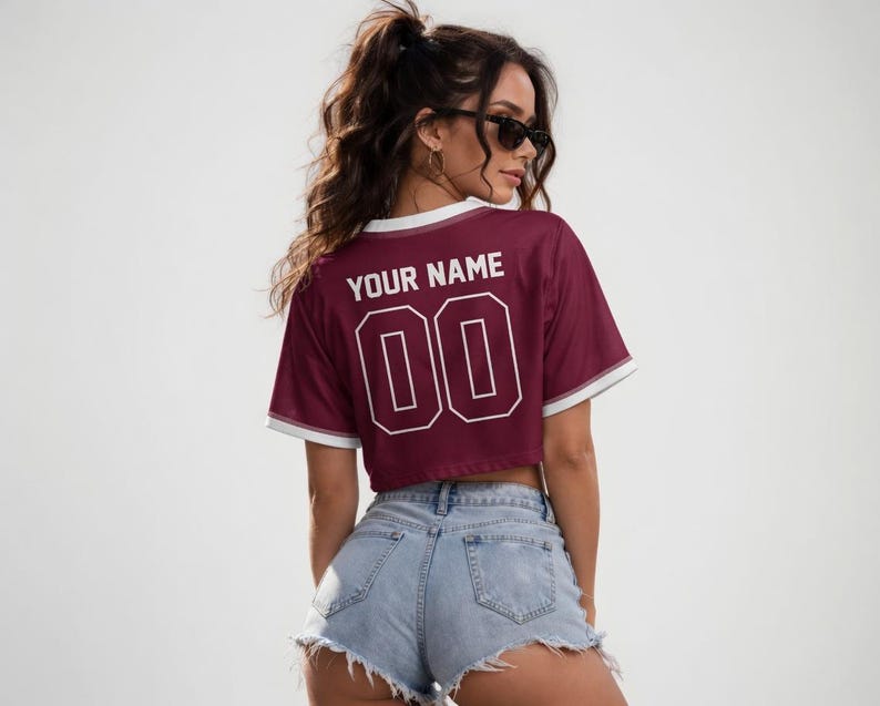 Peut inclure: Un crop top de couleur bordeaux avec des bordures blanches et le texte "YOUR NAME" et le num&eacute;ro "00" en blanc. Le maillot est associ&eacute; &agrave; un short en jean.
