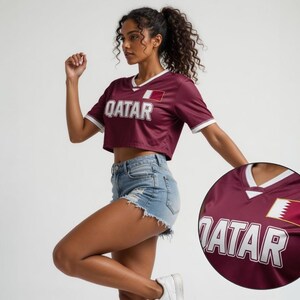 Peut inclure: Un maillot de corps bordeaux avec des bordures blanches et le mot "QATAR" en lettres majuscules blanches. Le maillot porte un &eacute;cusson du drapeau du Qatar sur la poitrine. Le mod&egrave;le porte un short en jean et des baskets blanches.