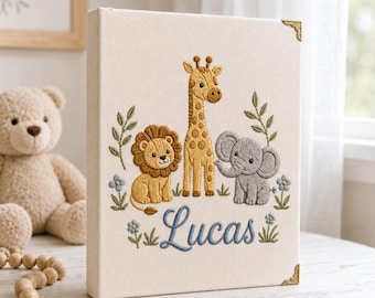 May include: A cream-colored fabric-covered photo album featuring embroidered illustrations of a lion, a giraffe, and an elephant. The name Lucas is embroidered in blue script below the animals, accented by small green leaves and blue flowers.