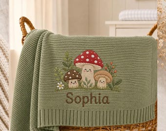 May include: A sage green knitted blanket featuring an embroidered design of three smiling mushrooms with a red and white spotted cap and two brown caps, surrounded by green foliage and small flowers, with the name Sophia stitched in brown thread.