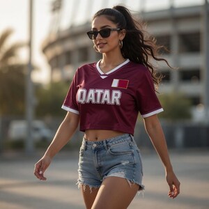 Peut inclure: Un maillot de corps bordeaux avec des bordures blanches et le mot "QATAR" en lettres majuscules blanches. Le maillot comporte &eacute;galement un petit embl&egrave;me de drapeau. La personne porte un short en jean et des lunettes de soleil.