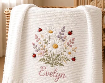 May include: A white knitted blanket featuring an embroidered bouquet of daisies, lavender, and small pink flowers, accented by four red ladybirds. The name Evelyn is embroidered in pink script lettering below the floral arrangement.