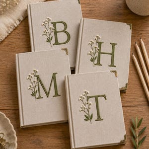May include: Four rectangular journals with beige fabric covers, each featuring an embroidered dark green letter and a delicate white floral sprig. Each journal has metallic gold-toned corner protectors on the front cover, displaying the letters B, H, M, and T.
