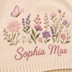 May include: A cream-coloured knit fabric featuring detailed embroidery of a pink butterfly and various wildflowers in shades of pink, purple, and white. The name Sophia Mae is embroidered in a cursive pink script font below the floral design.
