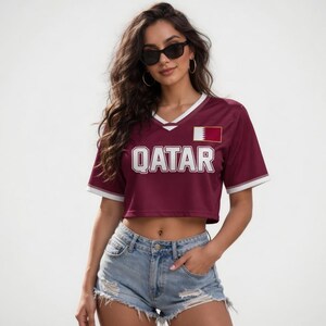 Peut inclure: Un crop top bordeaux avec des bordures blanches et le mot "QATAR" en lettres blanches. Le haut pr&eacute;sente un &eacute;cusson du drapeau du Qatar sur la poitrine droite. Le mod&egrave;le porte un short en jean et des lunettes de soleil.