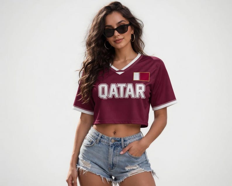 Peut inclure: Un crop top bordeaux avec des bordures blanches et le mot "QATAR" en lettres blanches. Le haut pr&eacute;sente un &eacute;cusson du drapeau du Qatar sur la poitrine droite. Le mod&egrave;le porte un short en jean et des lunettes de soleil.