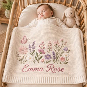May include: A cream-coloured knit baby blanket featuring an embroidered design of colourful wildflowers, a small pink butterfly, and the name Emma Rose stitched in pink cursive script. The blanket has a textured knit pattern and a ribbed border.