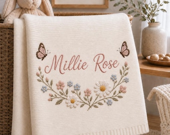 May include: A cream-coloured knit blanket featuring an embroidered floral wreath with daisies and small blue and pink flowers. The name Millie Rose is stitched in pink cursive script above the wreath, flanked by two brown and pink butterflies.