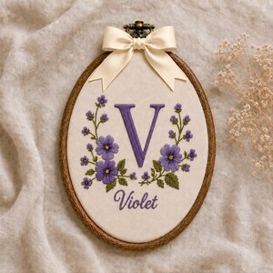 Puede incluir: Un bastidor de bordado ovalado de madera con tela color hueso, que presenta una letra V bordada en morado, flores moradas con hojas verdes y el nombre Violet en letra cursiva morada. Un lazo de satén color crema está sujeto en la parte superior.