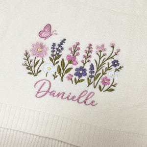 May include: An off-white textile featuring an embroidered design of various wildflowers in shades of pink, purple, and white, accompanied by a small pink butterfly. The name Danielle is embroidered in a cursive pink script below the flowers.